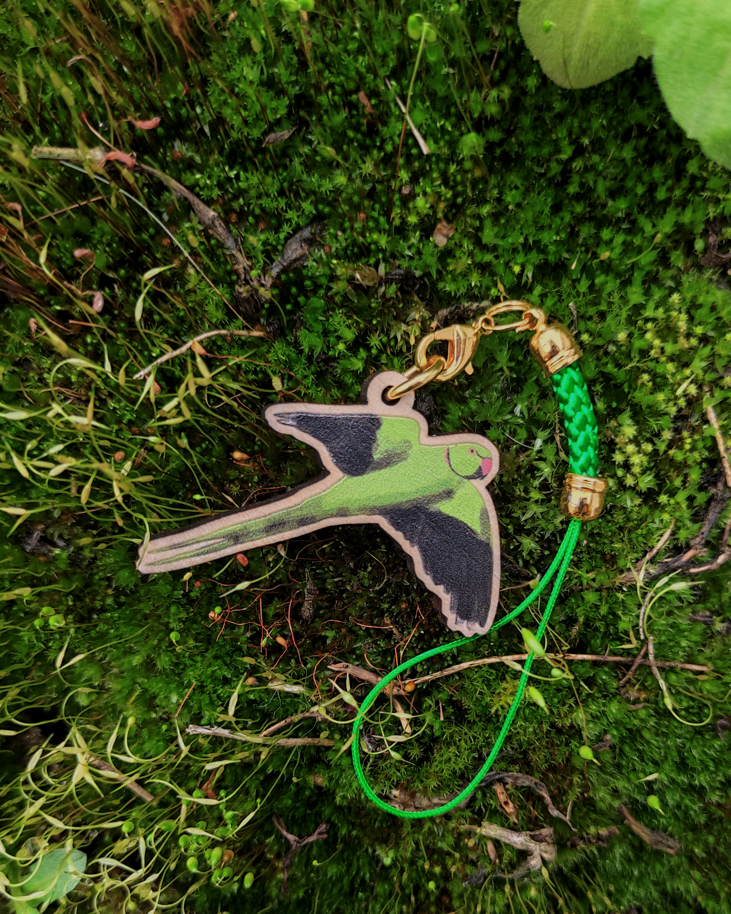 A wooden charm of a rose-ringed parakeet on green moss.