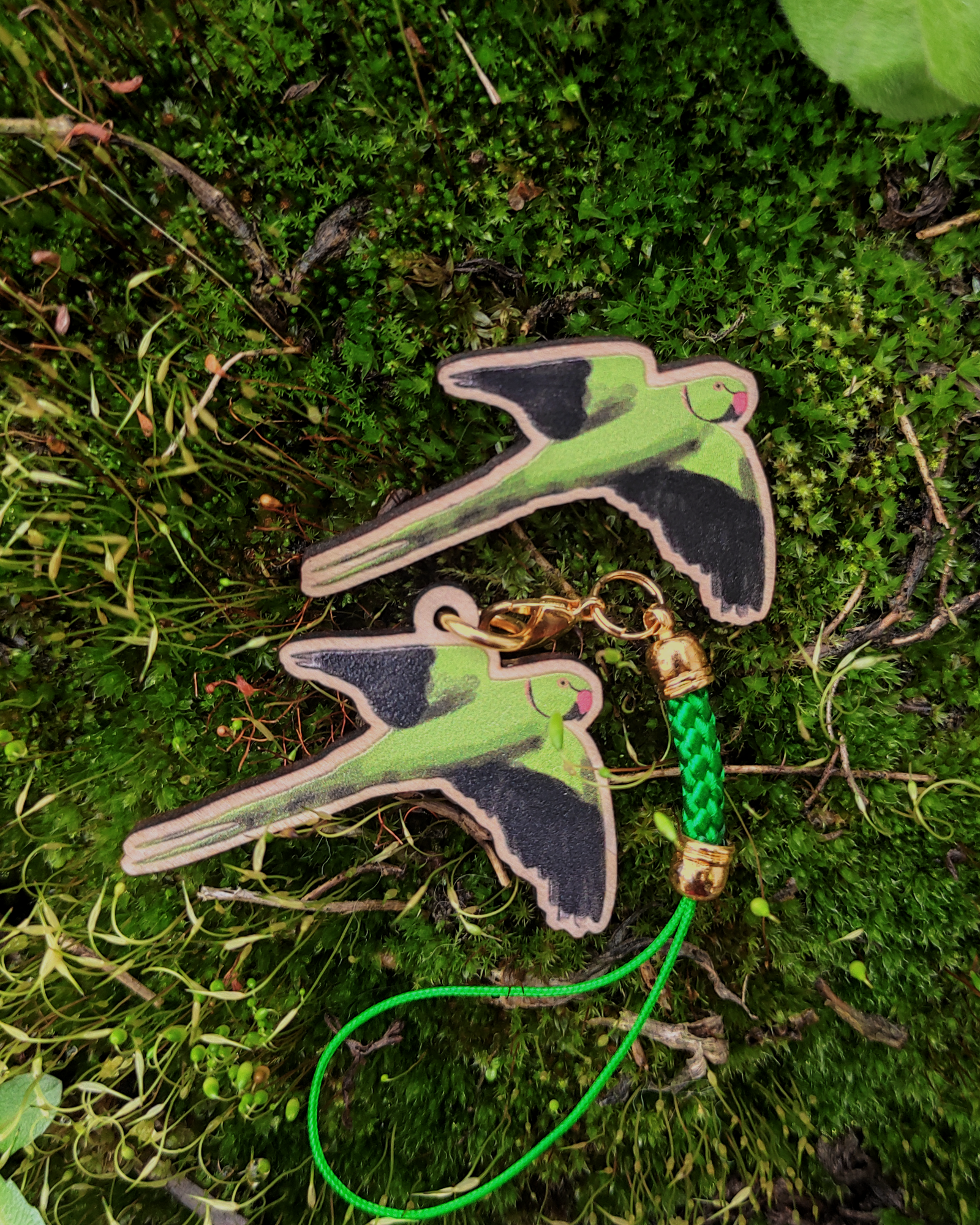 A wooden pin and charm of a rose-ringed parakeet on green moss.