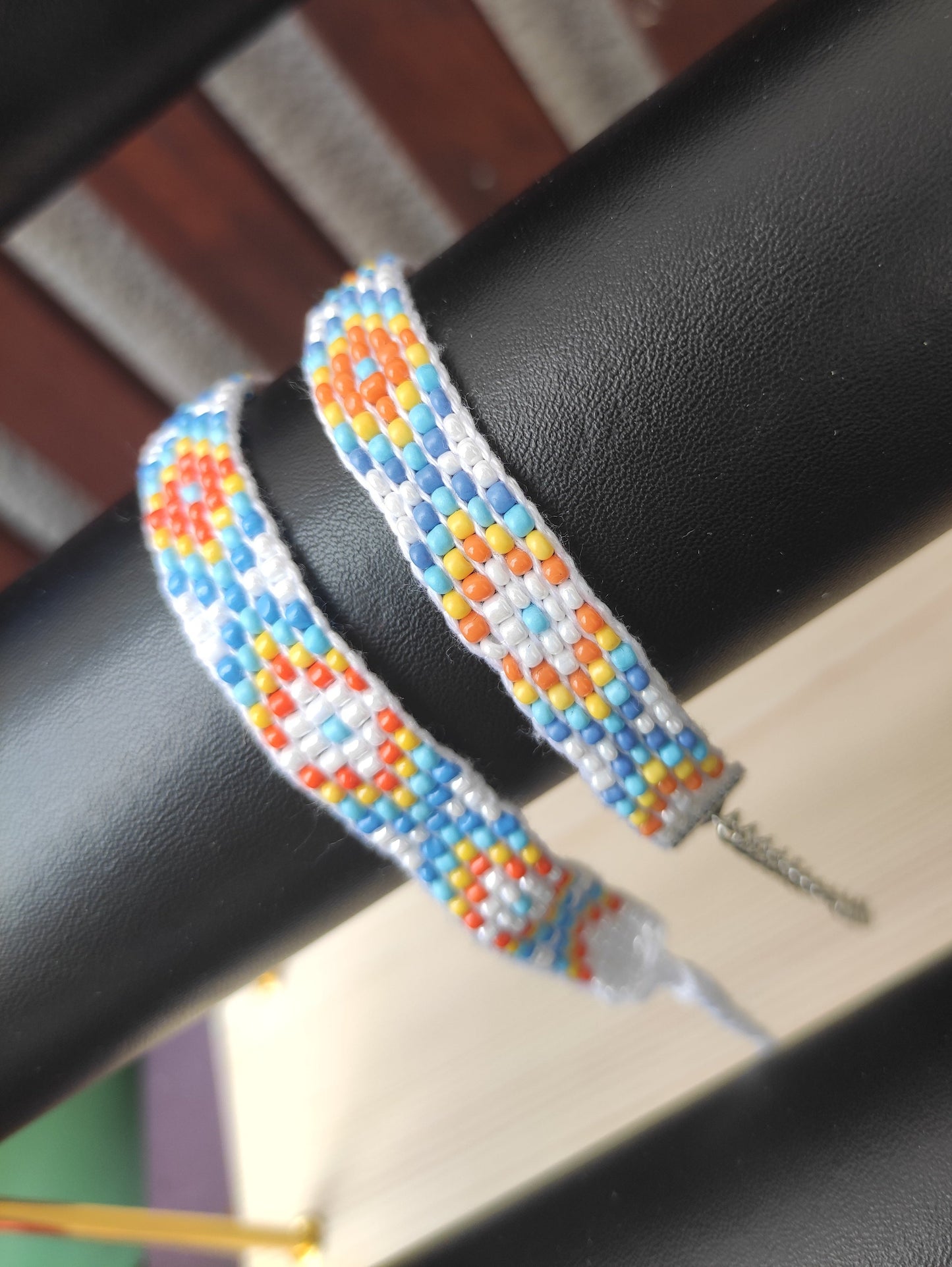 At the center of the picture are two bracelets on a display. They are made of rocaille pearls, which form a diagonal pattern. They are entirely made up of the colors that form the aroace flag (white, yellow, orange, sky blue, and marine).